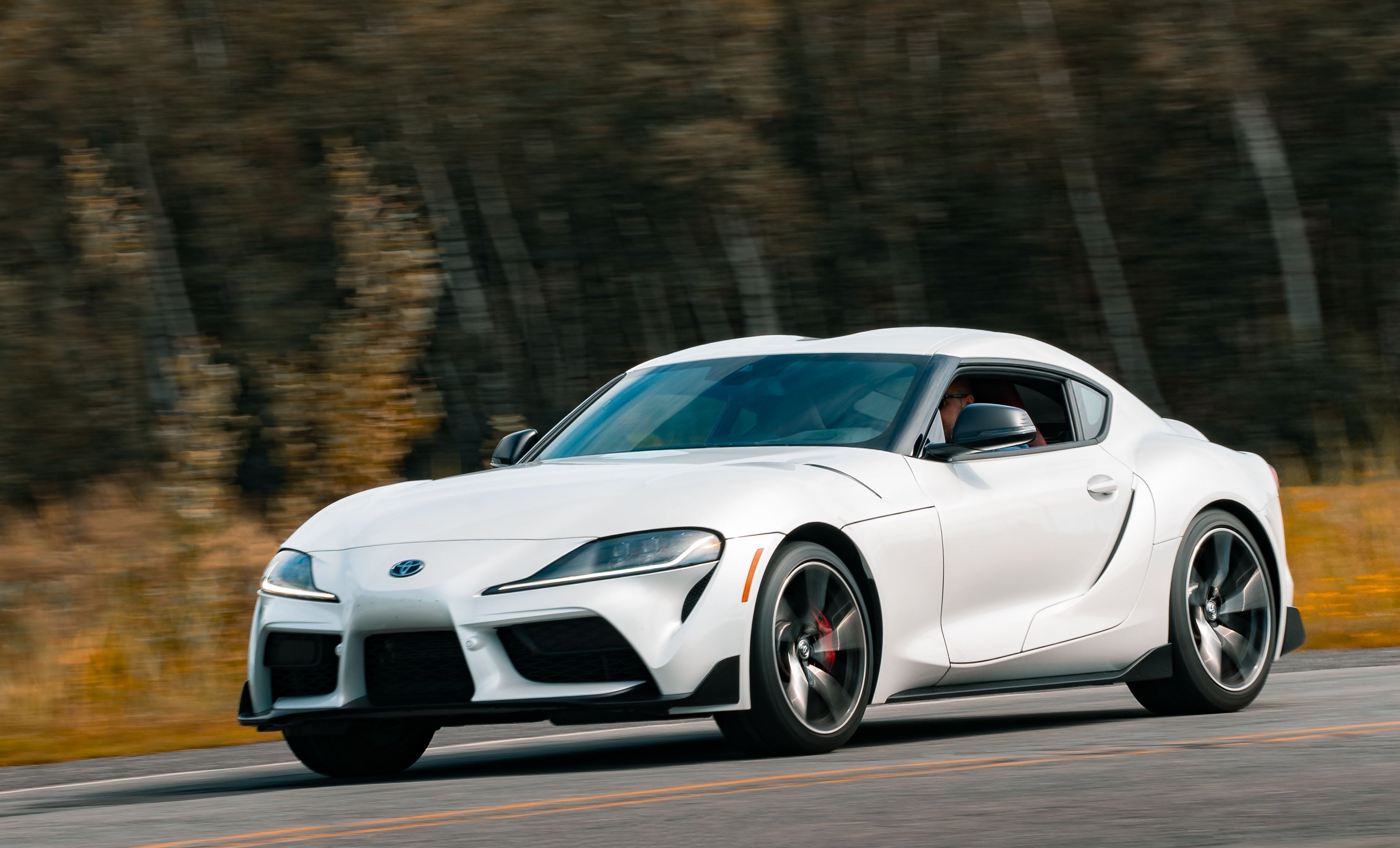 White 2020 Toyota Supra sports car driving on a road with blurred trees in the background
